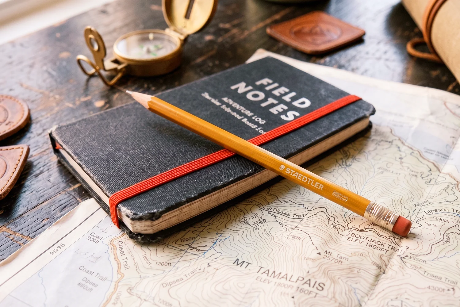 A worn field-notes journal and a pencil resting on a topographic trail map, beside an open brass compass.